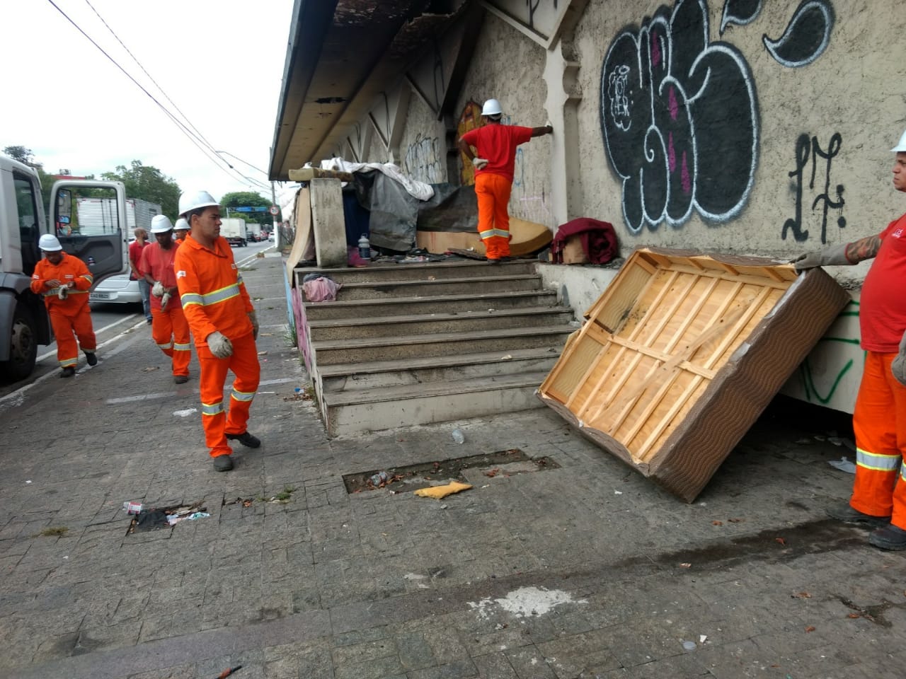 Foto: Cinco funcionário da Subprefeitura Ipiranga, vestidos com uniformas na cor laranja e capacetes brancos, todos estão recolhendo entulhos como colchões, papelão, restos de madeiras